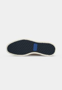 Semelle de chaussure avec grip bleu foncé à motif et bord extérieur blanc, montrant un logo rectangulaire bleu près de la zone de la voûte plantaire.