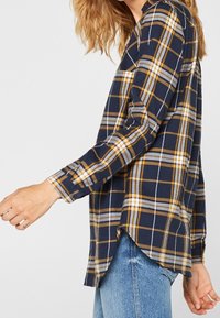 Flanelová košile s dlouhým rukávem v námořnické modři s kostkovaným vzorem ve žluté, šedé a bílé barvě. Má zaoblený lem a manžety na knoflíky. Měkká texturovaná látka.