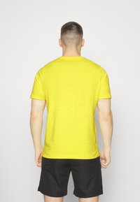 Camiseta de algodón amarilla con mangas cortas, cuello redondo y un corte relajado. Lleva unos pantalones cortos negros, vista desde la parte de atrás.