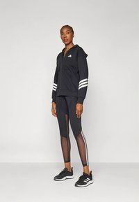 Camisola preta com fecho de correr e riscas brancas nas mangas, combinada com leggings pretas com painéis transparentes e riscas brancas laterais. Tênis desportivos pretos.
