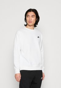 Valkoinen huppari, joka on valmistettu pehmeästä kankaasta ja jossa on pyöreä pääntie sekä pitkät hihat. Musta Nike-logo vasemmassa rinnassa. Yhdistetty tummiin housuihin.