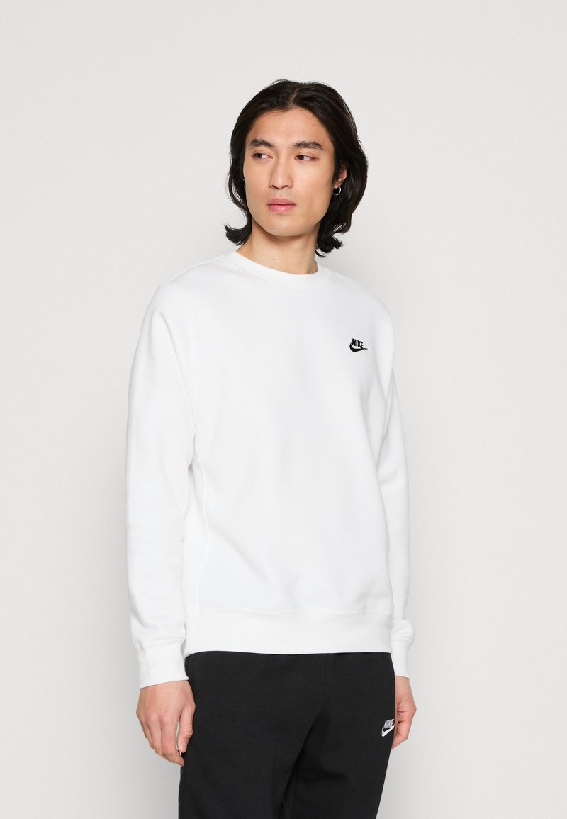 Sudadera blanca hecha de tela suave, con cuello redondo y mangas largas. Logo negro de Nike en el lado izquierdo del pecho. Combinada con pantalones oscuros.
