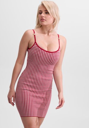 Blondynka w dopasowanej czerwono-białej sukience w kratę z cienkimi czerwonymi ramiączkami i gwiazdkowymi detalami na białym tle.
