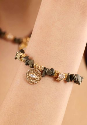 Armband aus dunklen grünen Steinplättchen, goldenen Akzenten und einem dekorativen goldenen Anhänger mit einem klaren Edelstein, der verschiedene Texturen und Formen aufweist.