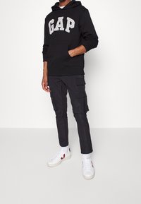 Black GAP hoodie with large gray embroidered logo, paired with black cargo pants featuring multiple pockets, and white sneakers with red accents.