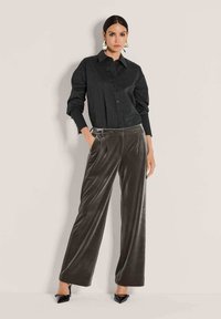 Vrouw die tegen een muur staat, gekleed in een zwarte knopenblouse, grijze wijdvallende fluwelen broek, zwarte puntige hakken en geometrische oorbellen.