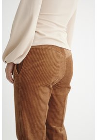 Calças de sarja castanhas com riscas verticais, apresentando bolsos laterais e uma cintura moldada. A parte de cima é uma camisa de manga longa de cor creme.