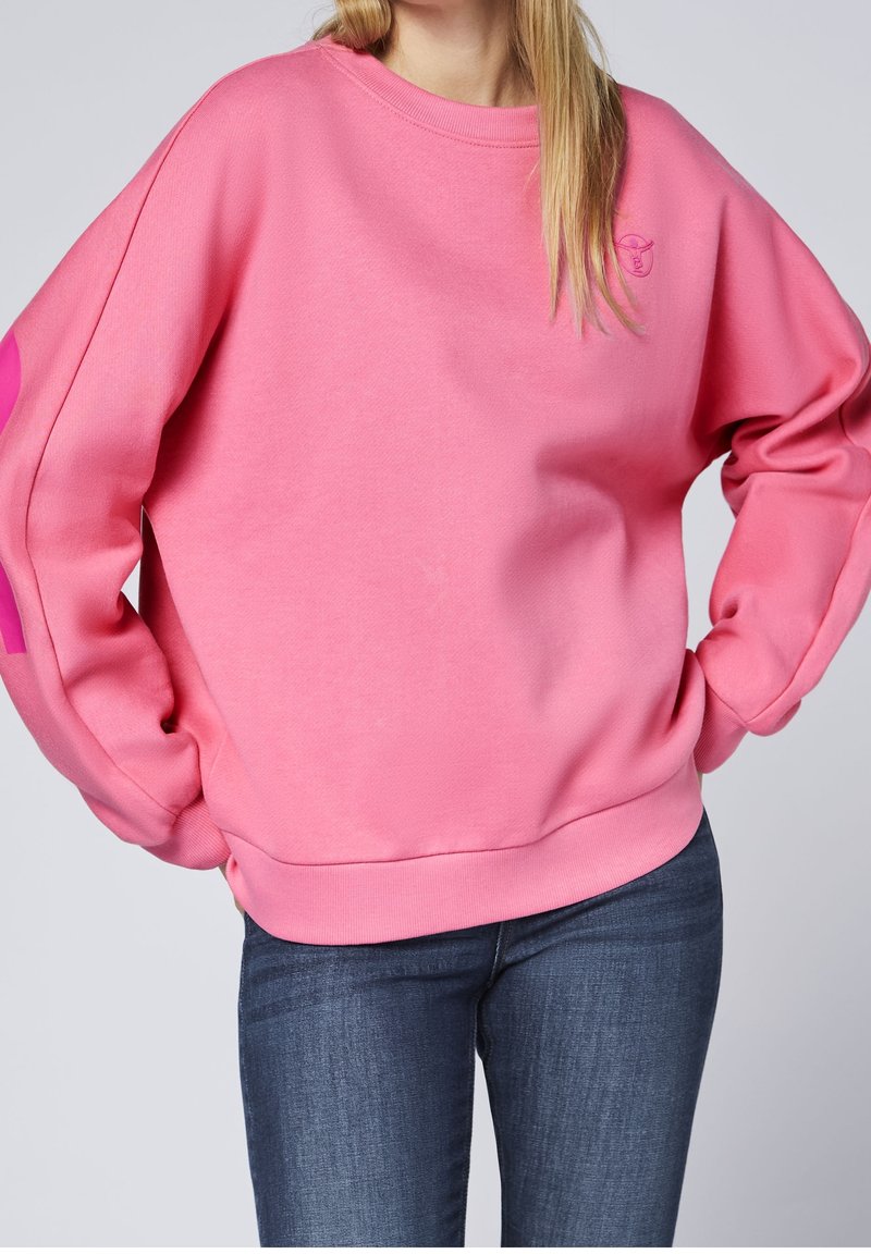 Femme portant un sweat-shirt rose ample avec un petit logo brodé sur la poitrine et un jean bleu foncé, mains dans les poches.