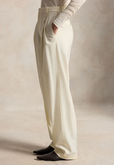 Φαρδιά παντελόνια σε ανοιχτό κίτρινο χρώμα από απαλό ύφασμα, με πιέτες μπροστά και πλαϊνές τσέπες, συνδυασμένα με μυτερές μαύρες παπούτσια.