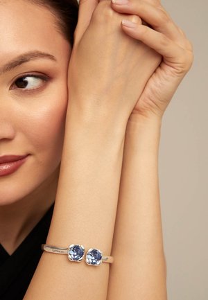 Primer plano de una persona que lleva una pulsera plateada con dos piedras preciosas azules, con los brazos cruzados cerca del rostro contra un fondo neutro.