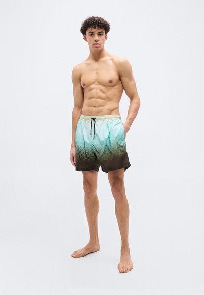 Νέος άνδρας στέκεται ξυπόλητος, φορώντας μαγιό με διαβάθμιση από τιρκουάζ σε καφέ, με σχεδιασμό μοτίβου και κορδόνι στη μέση.