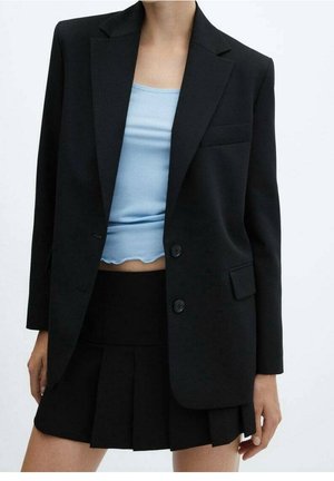 Kvinde iført en sort oversized blazer, sort plisseret mini nederdel og lyseblå ribbet crop top med bølget kant.