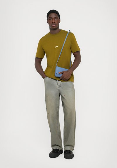 Man wearing olive green MSGM t-shirt, faded gray jeans, black shoes, and carrying a small blue crossbody bag, standing with one hand in pocket.