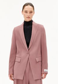 Helles rosa Blazer mit strukturiertem Design, Spitzenrevers, Taschen vorne und Knopfverschluss, getragen über einem schwarzen gerippten Rollkragenpullover.