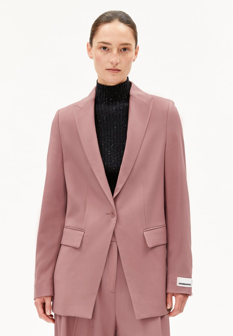 Helles rosa Blazer mit strukturiertem Design, Spitzenrevers, Taschen vorne und Knopfverschluss, getragen über einem schwarzen gerippten Rollkragenpullover.