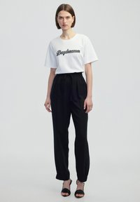 Witte t-shirt met korte mouwen met "Daydreamer" in zwarte borduursel, gecombineerd met zwarte, taps toelopende broek met elastische boorden en een strikband. Zwarte sandalen.