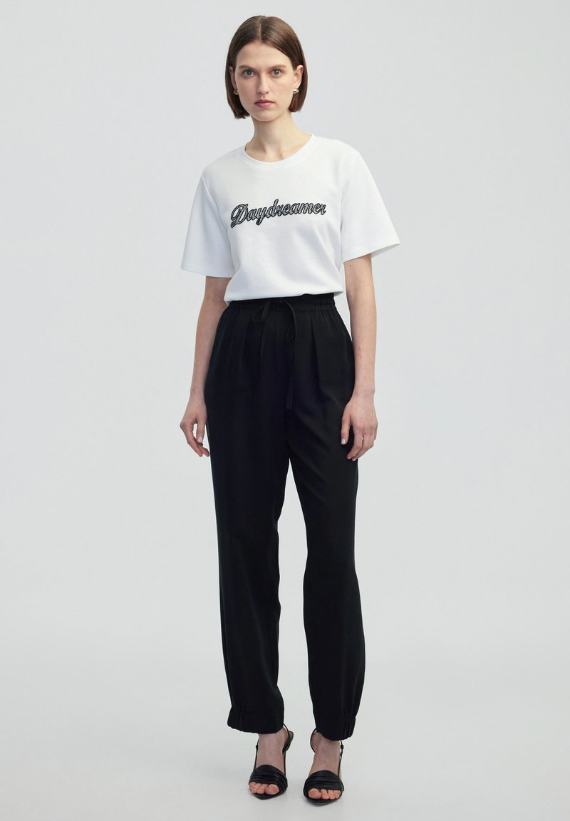 Witte t-shirt met korte mouwen met "Daydreamer" in zwarte borduursel, gecombineerd met zwarte, taps toelopende broek met elastische boorden en een strikband. Zwarte sandalen.