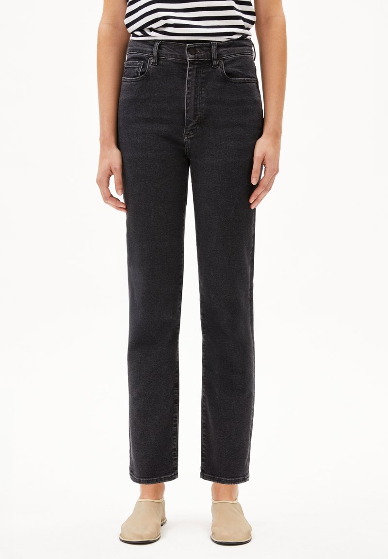Personne portant un jean noir taille haute à jambes droites, une chemise rayée noire et blanche, et des chaussures beige à enfiler, debout devant un fond blanc.