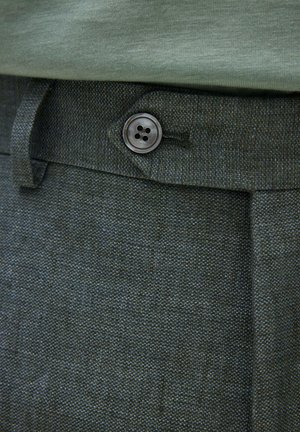 Primo piano di pantaloni scuri dalla texture evidenziata con un bottone nero a quattro fori e passante per cintura, abbinati a una camicia verde chiaro infilata dentro.