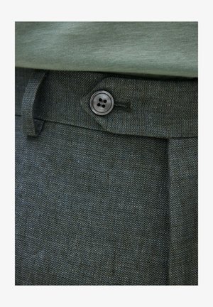 Gros plan sur un pantalon texturé foncé avec un bouton noir à quatre trous et une boucle de ceinture, associé à une chemise vert clair rentrée.