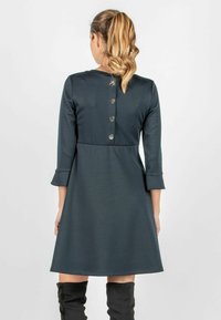 Dunkelgrünes Kleid mit Dreiviertelärmeln, tailliertem Oberteil, ausgestelltem Rock und fünf metallischen Knöpfen am Rücken. Glatte Stoffstruktur.