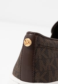 Chaussure en cuir marron avec motif texturé et accent en or du logo MK. Présente une semelle blanche et des détails cousus le long du bord.