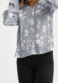 Blusa floreale blu chiaro con motivi bianchi, scollatura a V e maniche a palloncino. Indossata con jeans neri, con orlo curvo.