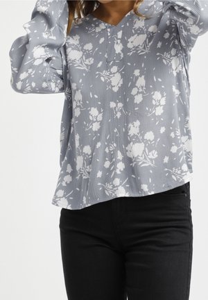 Hellbluse mit floralen Muster in Weiß, V-Ausschnitt und puffigen Ärmeln. Kombiniert mit schwarzen Jeans mit abgerundetem Saum.
