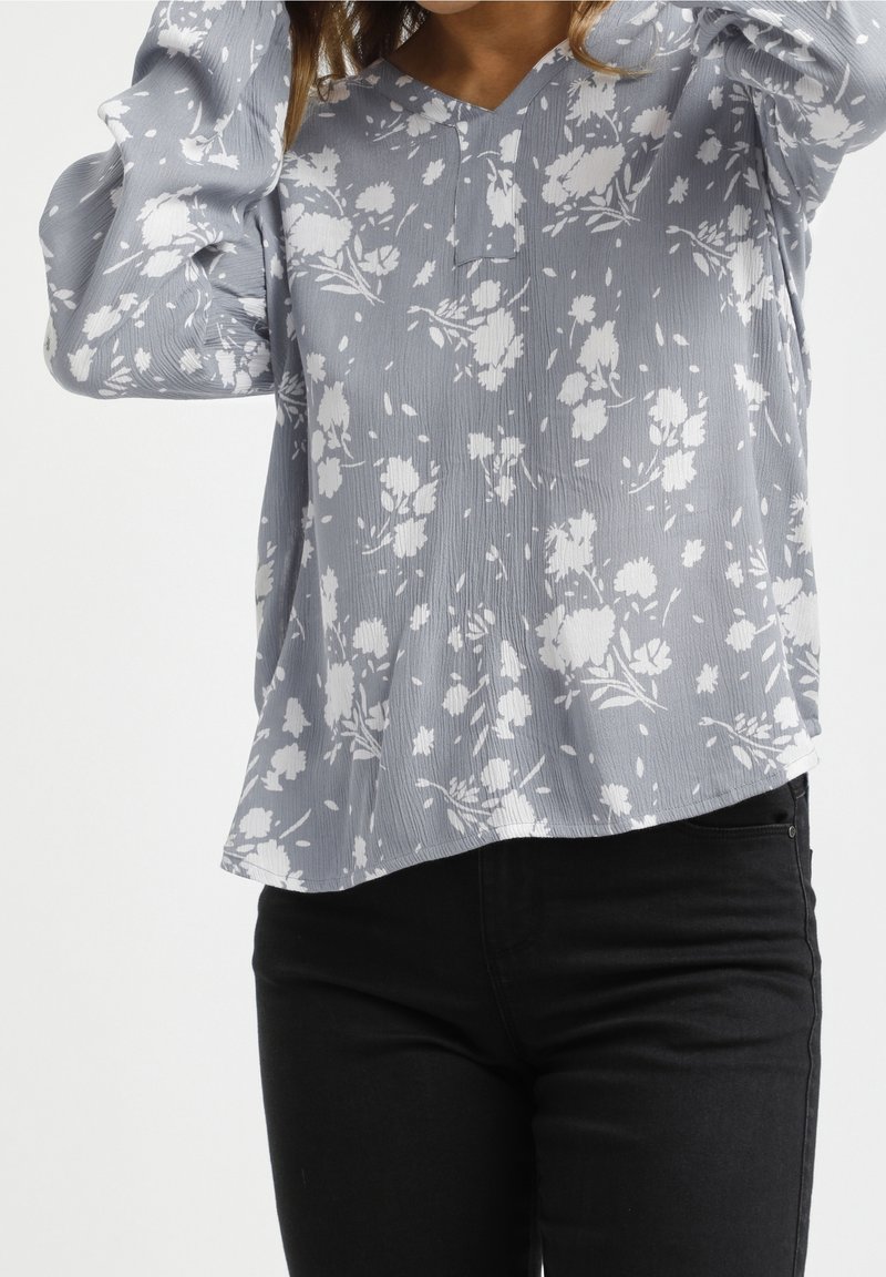 Blusa floreale blu chiaro con motivi bianchi, scollatura a V e maniche a palloncino. Indossata con jeans neri, con orlo curvo.