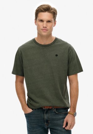 Mørkegrøn kortærmet T-shirt med rund hals, prydet med et lille sort logo på brystet, båret med blå jeans og et ur.