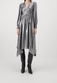 Robe argentée métallique à manches longues, décolleté en V, corsage plissé et ourlet asymétrique, associée à des bottes noires montantes.