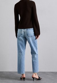 Brązowy dziergany top z rozkloszowanymi rękawami o fakturowanym wzorze; zestawiony z jasnoniebieskimi jeansami o prostych nogawkach oraz czarnymi sandałami na obcasie.