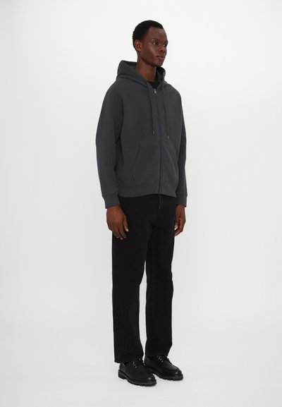 Dark gray zip-up hoodie with a spacious front pocket, drawstring hood, and ribbed cuffs, paired with black straight-leg pants and black shoes.