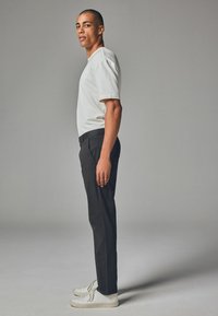 Männliches Model in einem taillierten, kurzärmeligen weißen Hemd, schwarzen Hosen und weißen Sneakers, seitlich vor einem neutralen grauen Hintergrund posierend.