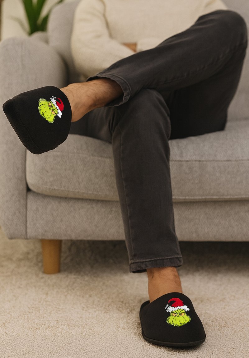 Black slippers with a thick sole feature an embroidered Grinch design in green, wearing a red Santa hat, on the front. Textured textile material.