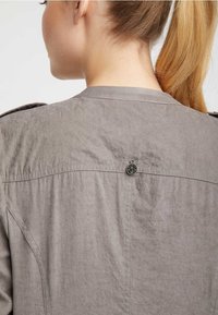 Camisa ligera gris con una textura ligeramente arrugada, que presenta un detalle de botón en la parte posterior y un diseño de cuello clásico.