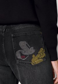 Jean en denim noir avec une texture délavée, présentant un design sur la poche arrière avec des graphismes de personnages de dessin animé gris et jaune sur un fond sombre.