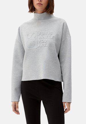 Kvinde iført grå langærmet turtleneck-trøje med hævet tekst "FUN IS WHERE YOU ARE" og sorte bukser, stående mod en hvid baggrund.
