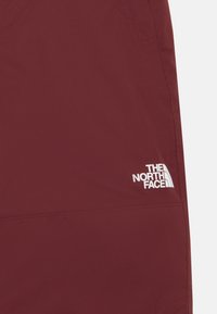 Bordové outdoorové nohavice s hladkou textúrou a bielym logom na dolnom pravom rohu. Vlastnosti zahŕňajú bočné vrecká a zosilnené švy.