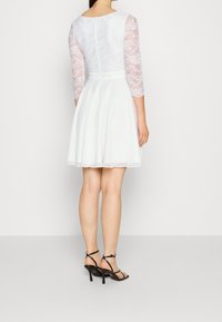 Weißes Kleid mit einem figurbetonten Spitzenoberteil und ausgestellt Rock, mit Dreiviertel-Spitzenärmeln und einem Reißverschluss im Rücken. Schwarze Riemchenabsätze.