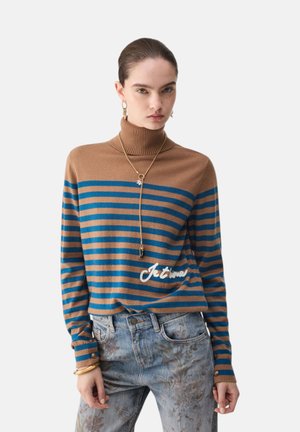 Donna che indossa un maglione a collo alto a righe marroni e blu con testo "Je t'aime", jeans con motivo floreale, collana d'oro, orecchini e braccialetti.