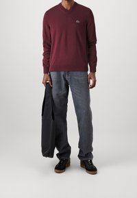 Pull en V bordeaux avec des poignets côtelés, porté sur un jean bleu clair. Une personne tient un sac noir et porte des baskets noires avec des semelles marron.