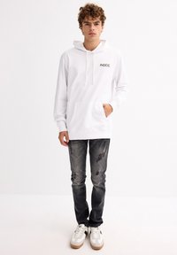 Sudadera blanca con capucha y la impresión "INSIDE" en el pecho, bolsillo delantero, combinada con jeans oscuros desgastados y zapatillas blancas con suelas beige.