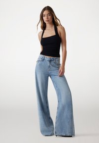 Jeans de tiro ancho en azul claro con un sutil brillo, combinados con una blusa sin mangas negra con escote cuadrado y textura fruncida.