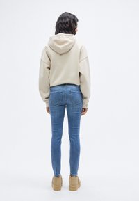 Μπεζ φούτερ με μεγάλο κουκούλι, φορεμένο πάνω από στενά μπλε τζιν. Τα παπούτσια είναι ταμπά με σόλα πλατφόρμας. Φαίνεται μακριά, σκούρα κυματιστά μαλλιά.