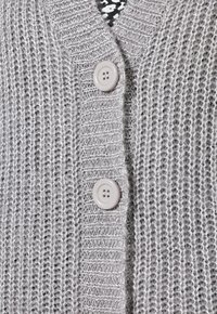 Grå stickad cardigan med v-ringning, försedd med två stora krämfärgade knappar och ett strukturerat mönster. Mjukt material med öppen väv.