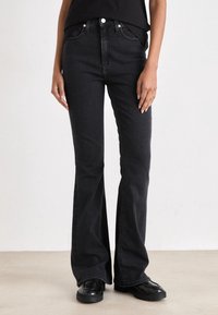 Svarta utsvängda jeans i denim med medelhög midja, fem fickor och metallbeslag. Har en slät textur och en åtsittande silhuett.