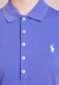 Lila polo skjorta i en strukturerad tyg; har en klassisk krage, fem knappar och en liten vit logotyp på vänster bröst.