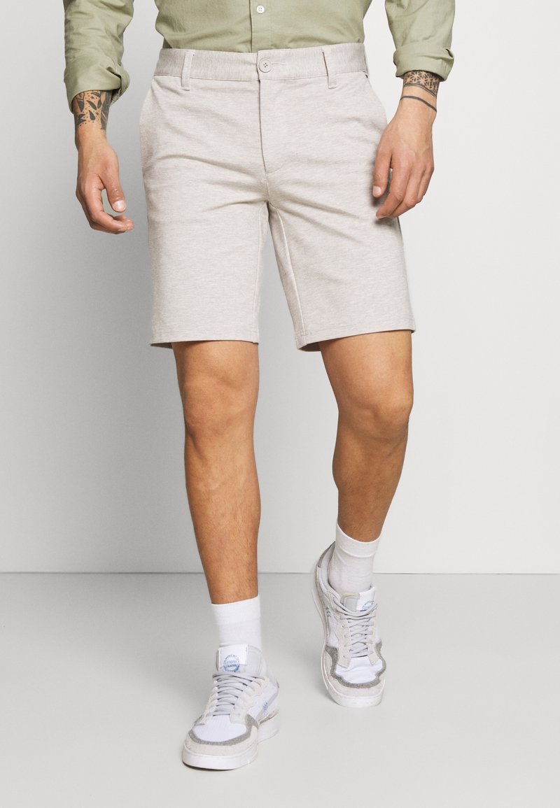 Homme portant un short beige longueur genou, des baskets blanches, des chaussettes blanches et une chemise vert clair à manches longues.