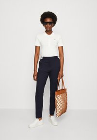 Polo bianco a maniche corte, pantaloni blu, scarpe slip-on bianche e una borsa a tracolla tessuta arancione. Occhiali da sole neri.